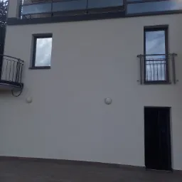 Nowoczesny budynek z balkonami o metalowych barierkach i dużymi oknami odbijającymi niebo, widok z zewnątrz na elewację w jasnym odcieniu szarości, ciemne płytki na podłożu.