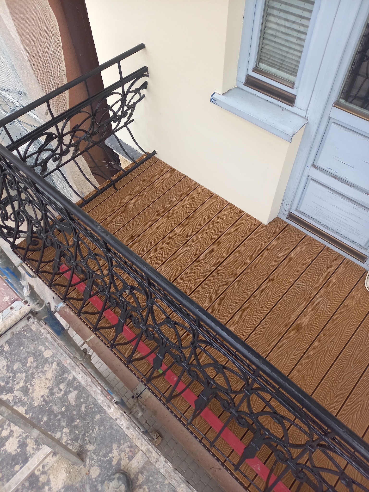 Balkon z nową, brązową podłogą z desek kompozytowych, czarna, ozdobna balustrada, widok z góry, fragment rusztowania w lewym dolnym rogu.
