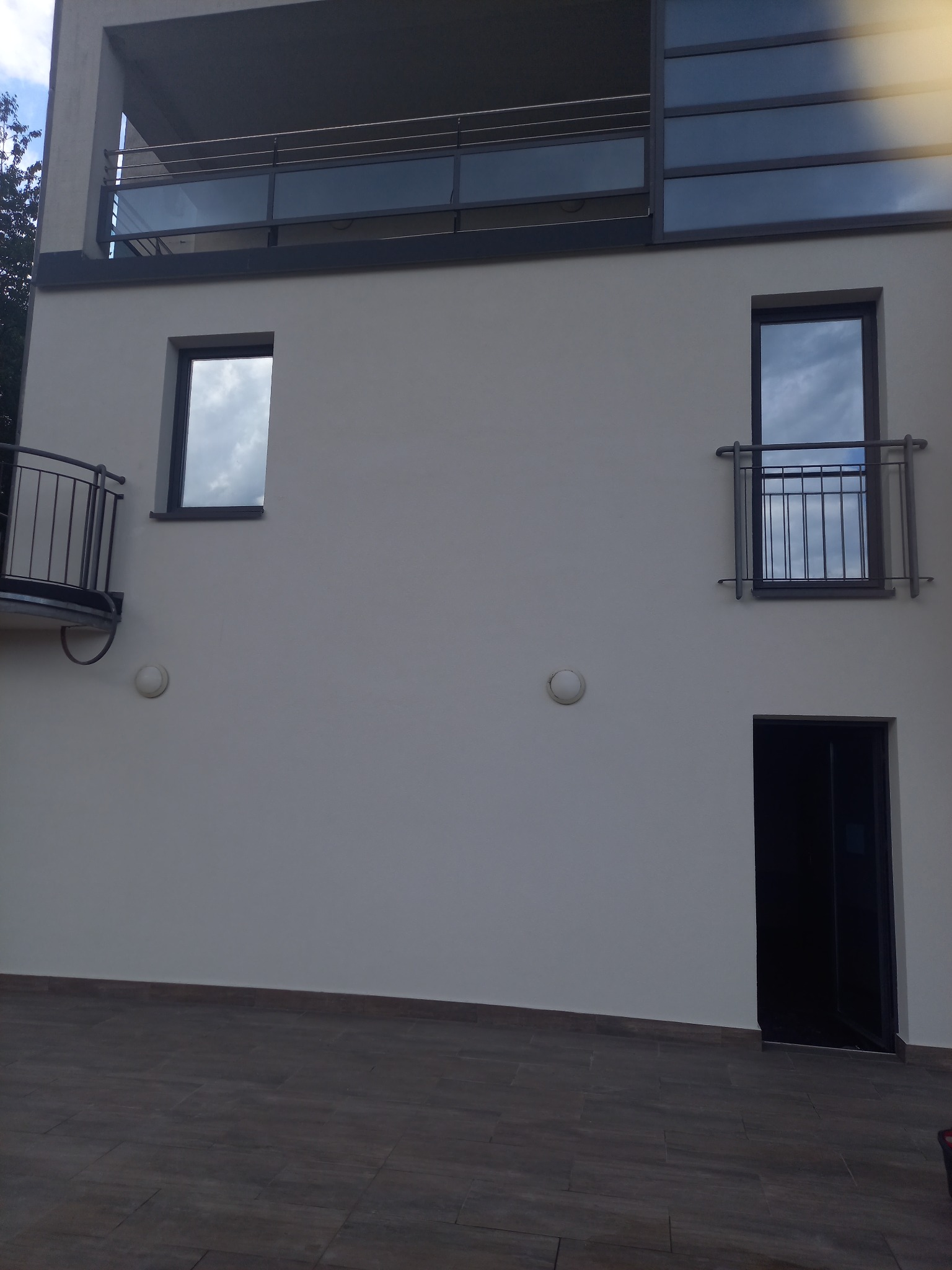 Nowoczesny budynek z balkonami o metalowych barierkach i dużymi oknami odbijającymi niebo, widok z zewnątrz na elewację w jasnym odcieniu szarości, ciemne płytki na podłożu.