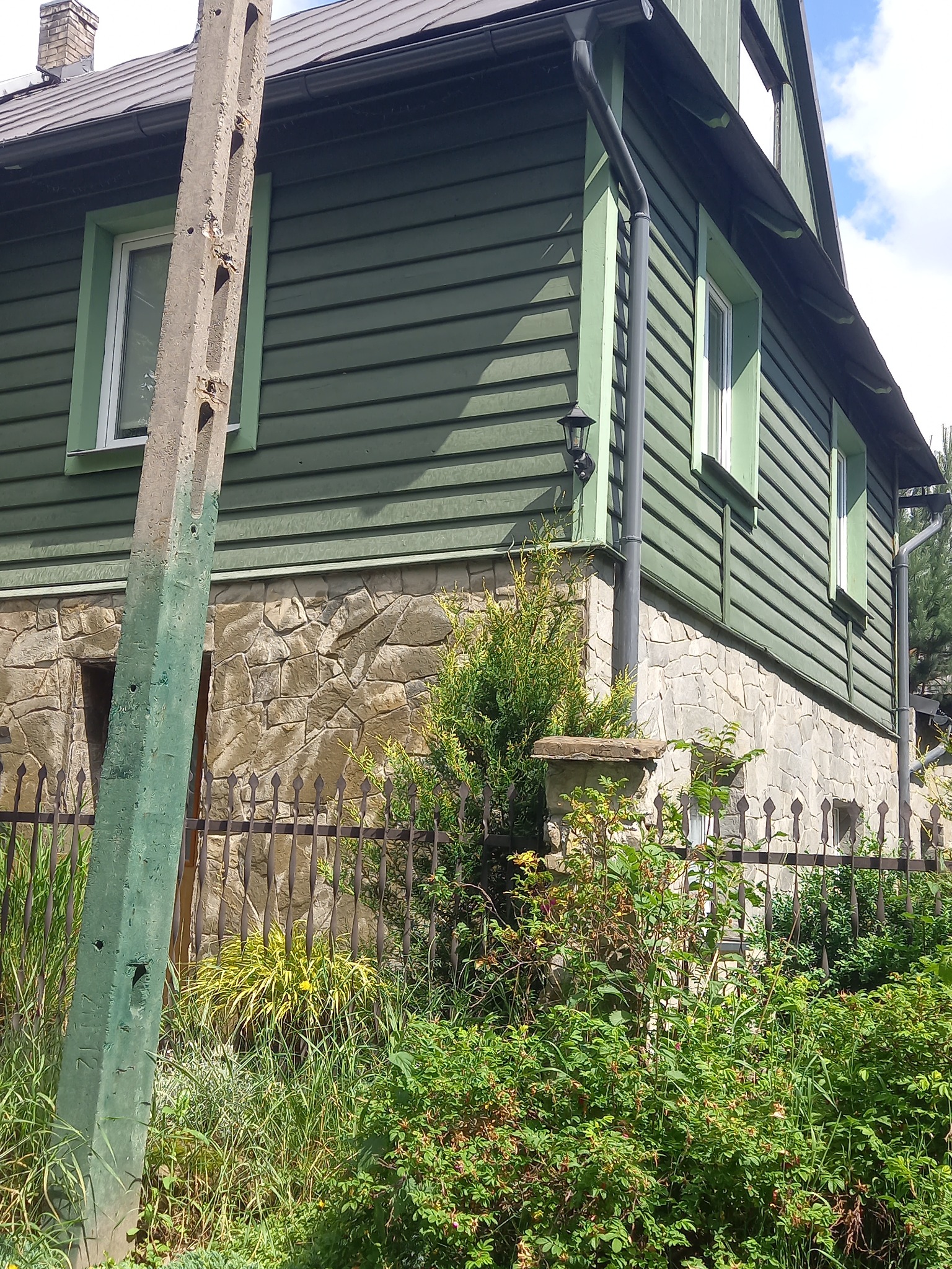 Zielony dom z drewnianą elewacją i kamienną podmurówką, widoczny zza metalowego ogrodzenia i bujnej zieleni, z betonowym słupem energetycznym na pierwszym planie.