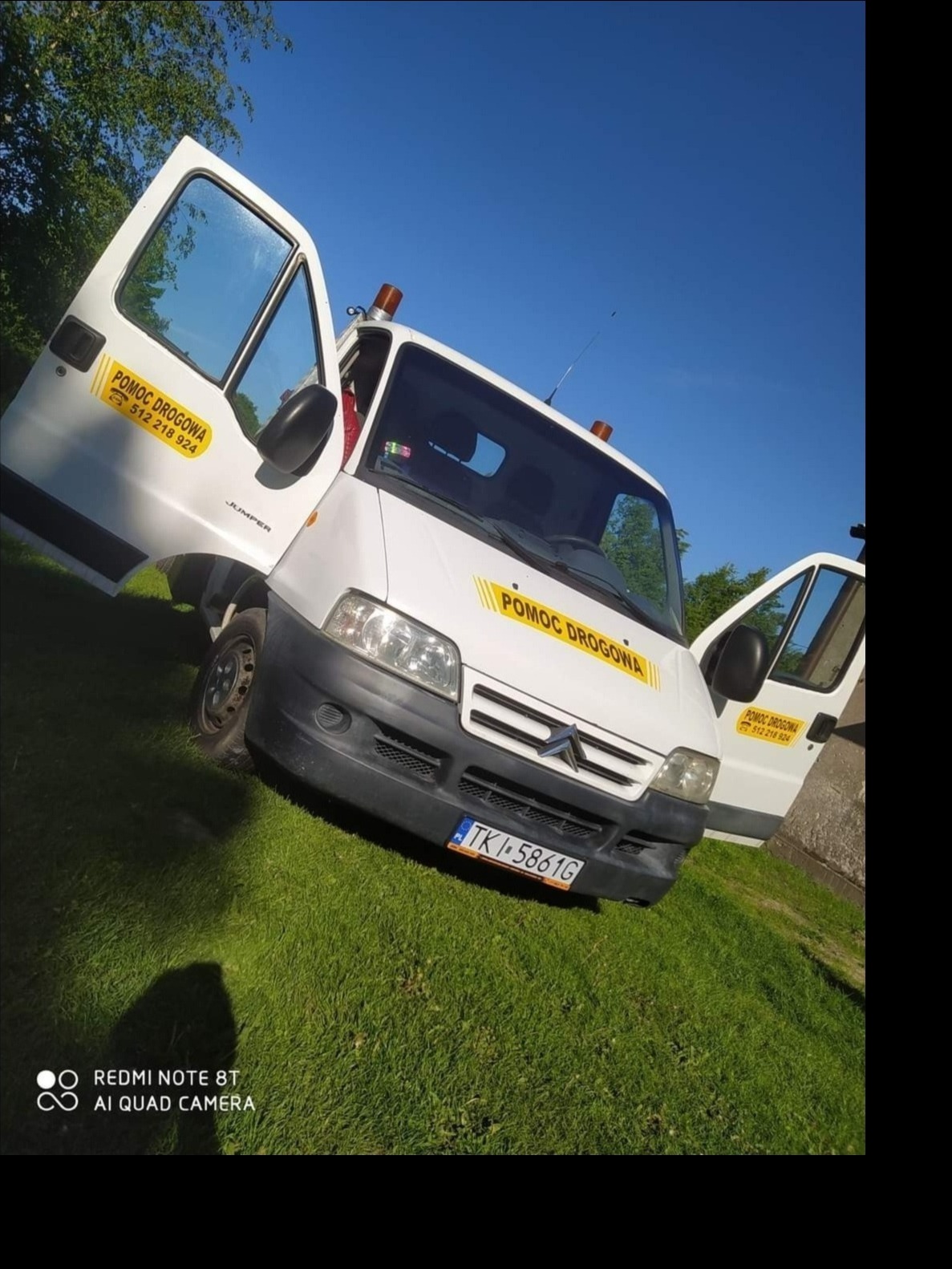Biała furgonetka z otwartymi drzwiami na trawniku, z napisem 'Pomoc Drogowa' i numerem telefonu na boku, widoczny numer rejestracyjny TKI 5861G.