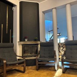 My Way - Ośrodek leczenia uzależnień - Wnętrze ośrodka: trzy fotele z drewnianymi ramami ustawione w przestronnym pokoju z kominkiem i dekoracyjną ścianą z pionowymi panelami w kolorze czerni i złota.