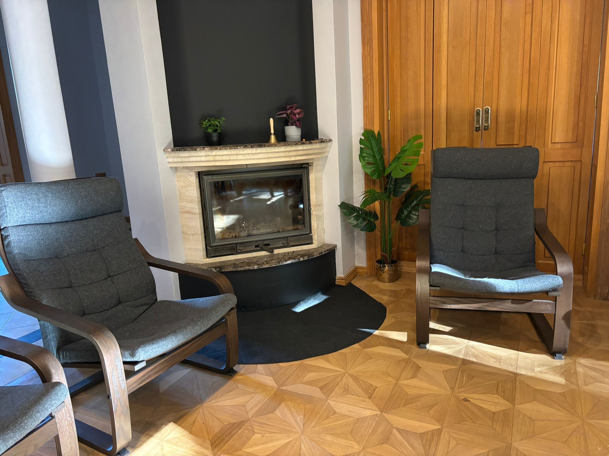 Przytulne wnętrze z dwoma fotelami, kominkiem z marmurowym obramowaniem i drewnianą podłogą w geometryczny wzór. Rośliny doniczkowe dodają naturalnego akcentu.
