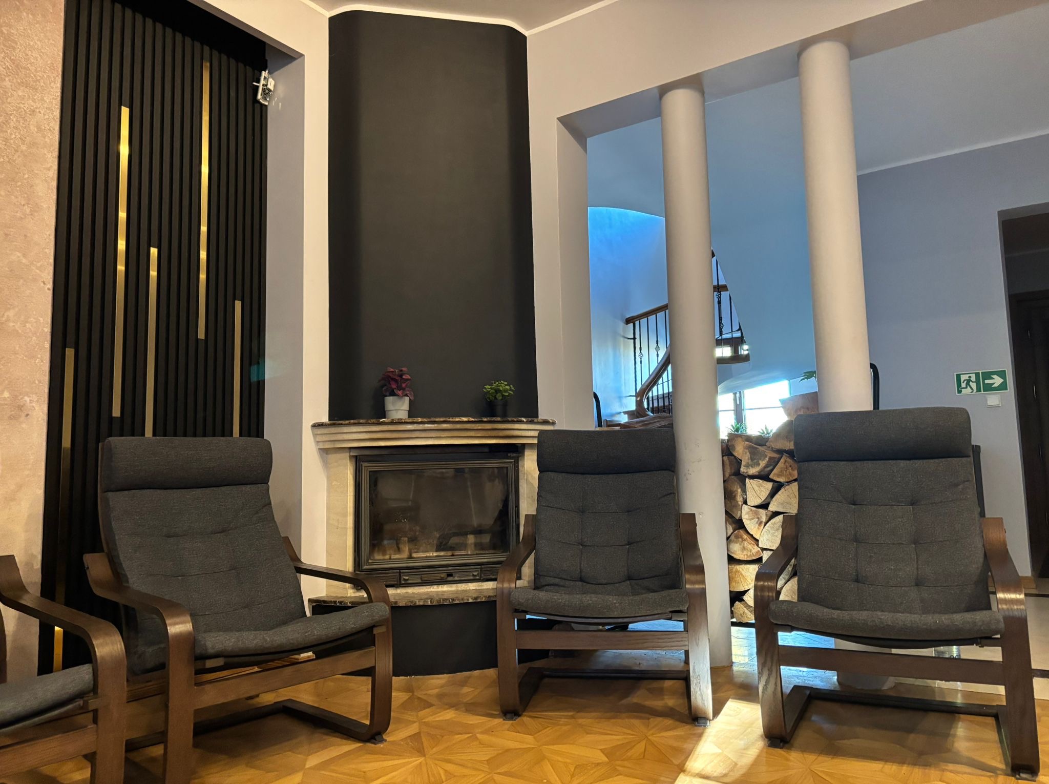 Wnętrze ośrodka: trzy fotele z drewnianymi ramami ustawione w przestronnym pokoju z kominkiem i dekoracyjną ścianą z pionowymi panelami w kolorze czerni i złota.