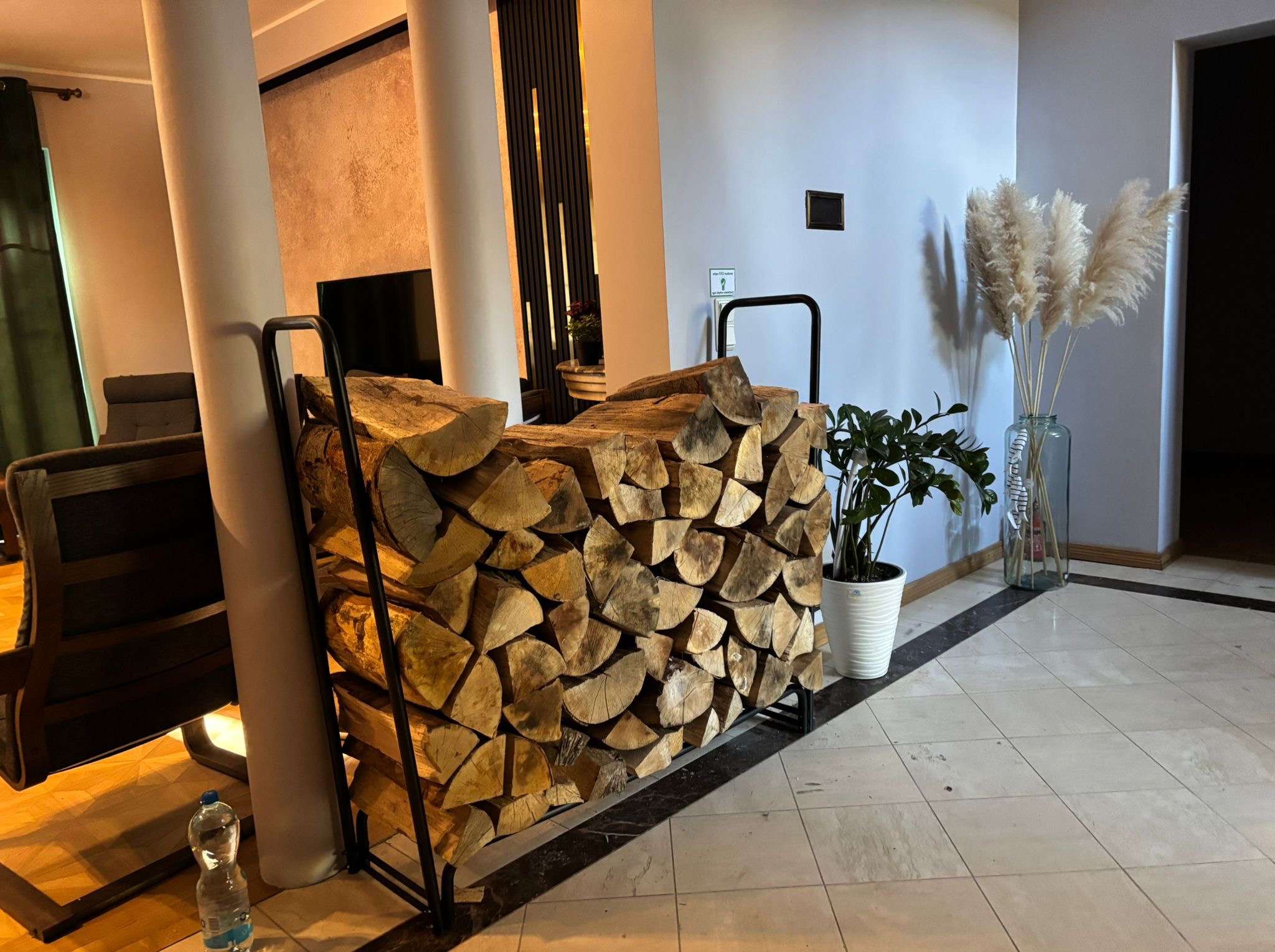 Drewno kominkowe ułożone w metalowym stojaku w nowoczesnym wnętrzu. Obok roślina w doniczce i trawa pampasowa w szklanym wazonie. W tle kolumny i telewizor.