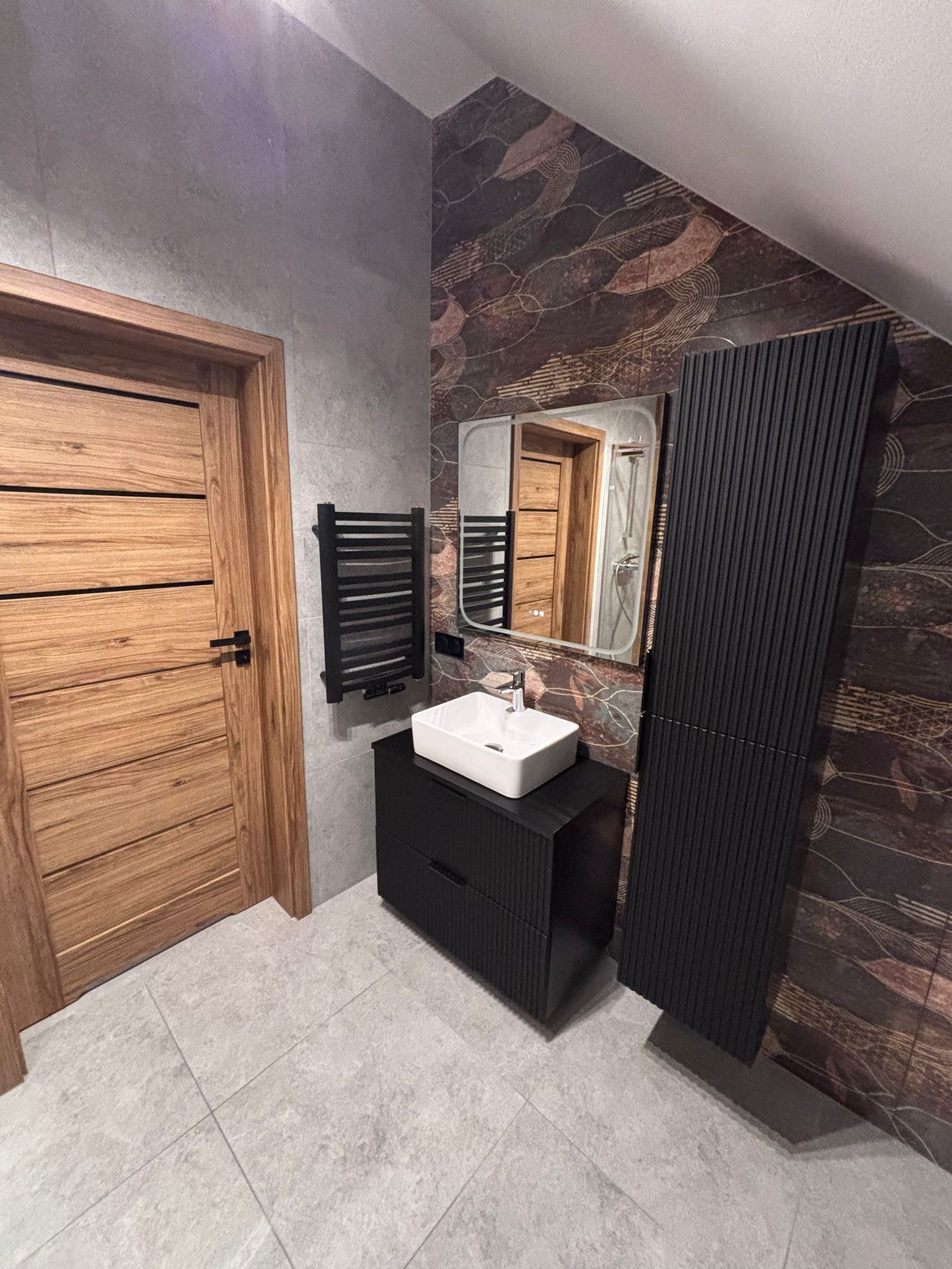 Nowoczesna łazienka na poddaszu: drewniane drzwi z poziomymi panelami, czarna szafka z umywalką, grzejnik drabinkowy i dekoracyjna ściana z motywem liści.