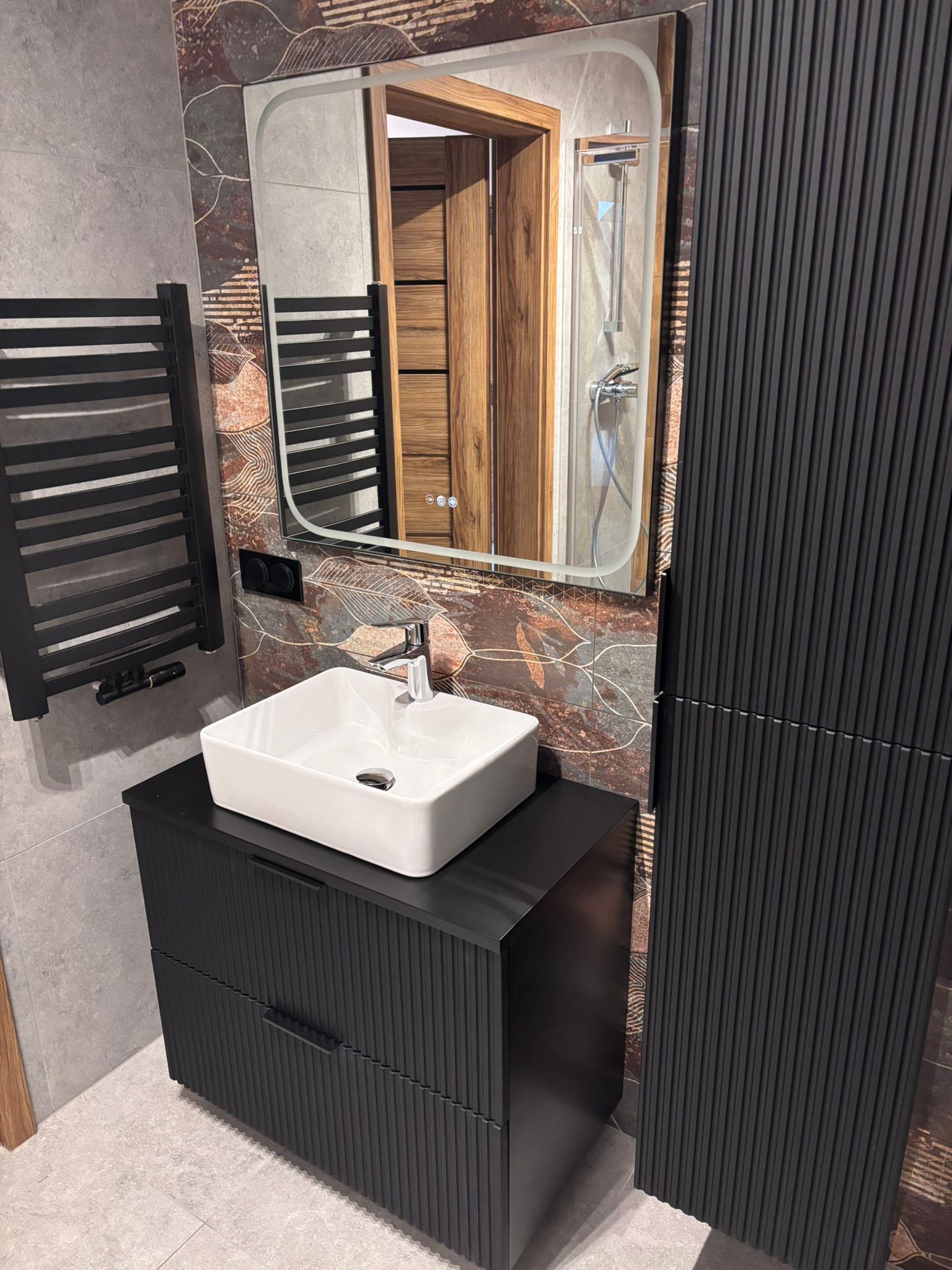 Nowoczesna łazienka w Kąpinie z czarną szafką umywalkową, białym blatem i lustrem. Widoczny grzejnik drabinkowy i dekoracyjne płytki na ścianie.