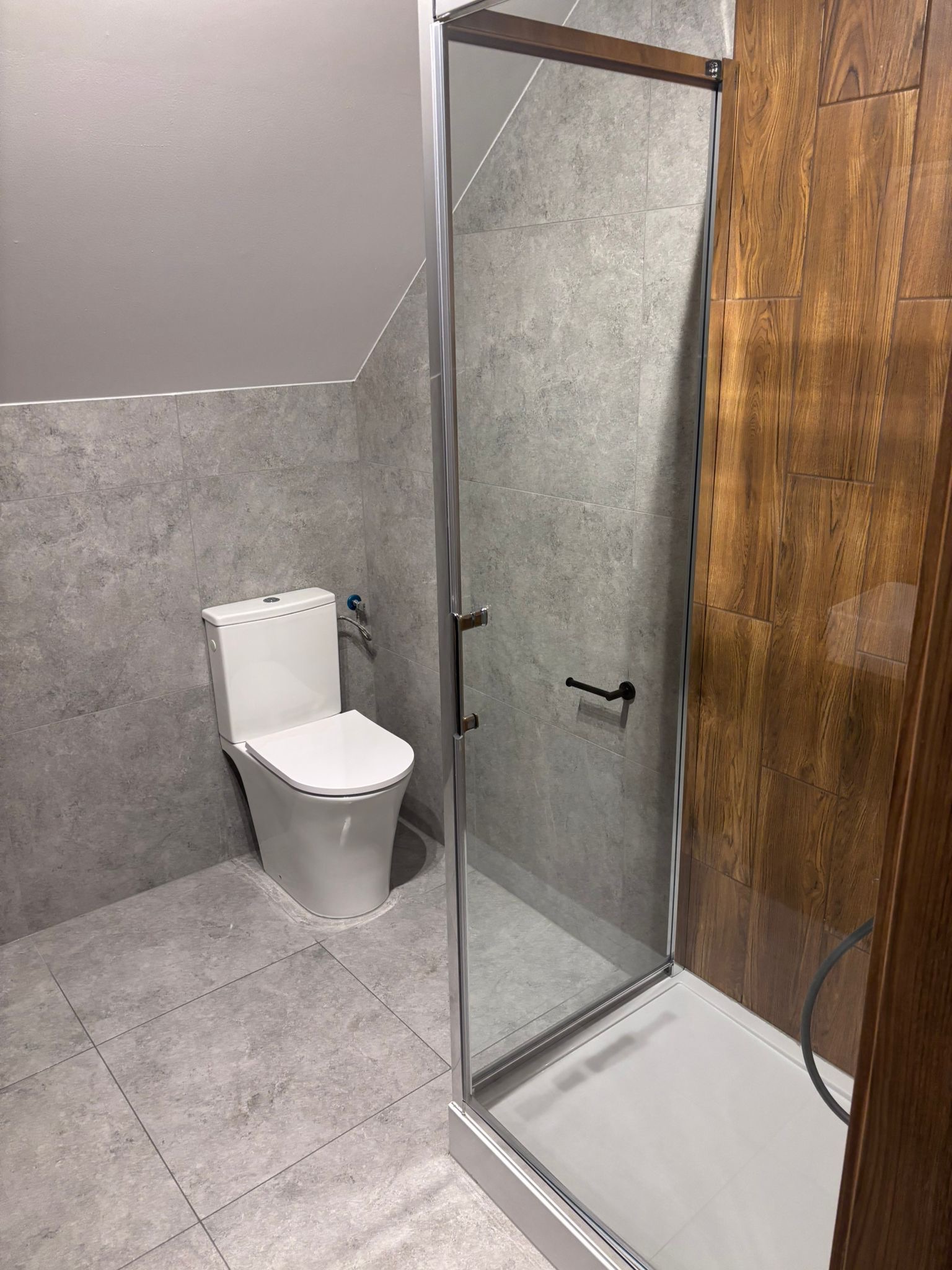 Nowoczesna łazienka po remoncie: szare płytki, biała toaleta i kabina prysznicowa z drewnianymi akcentami. Widok na ukończone wnętrze z bliska.