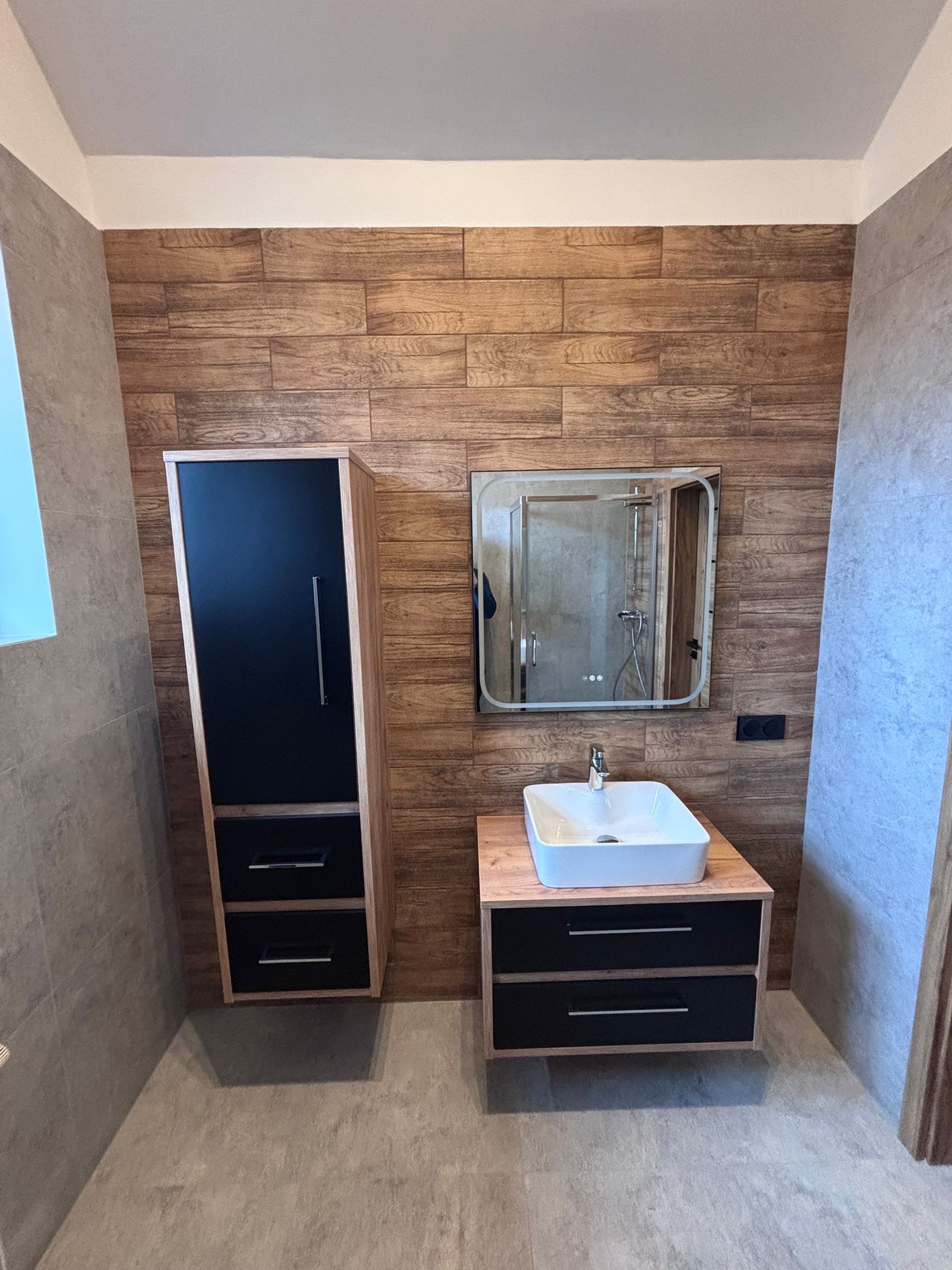 Nowoczesna łazienka z drewnianą ścianą, szafką z czarnymi frontami i umywalką nablatową. W lustrze odbija się kabina prysznicowa.