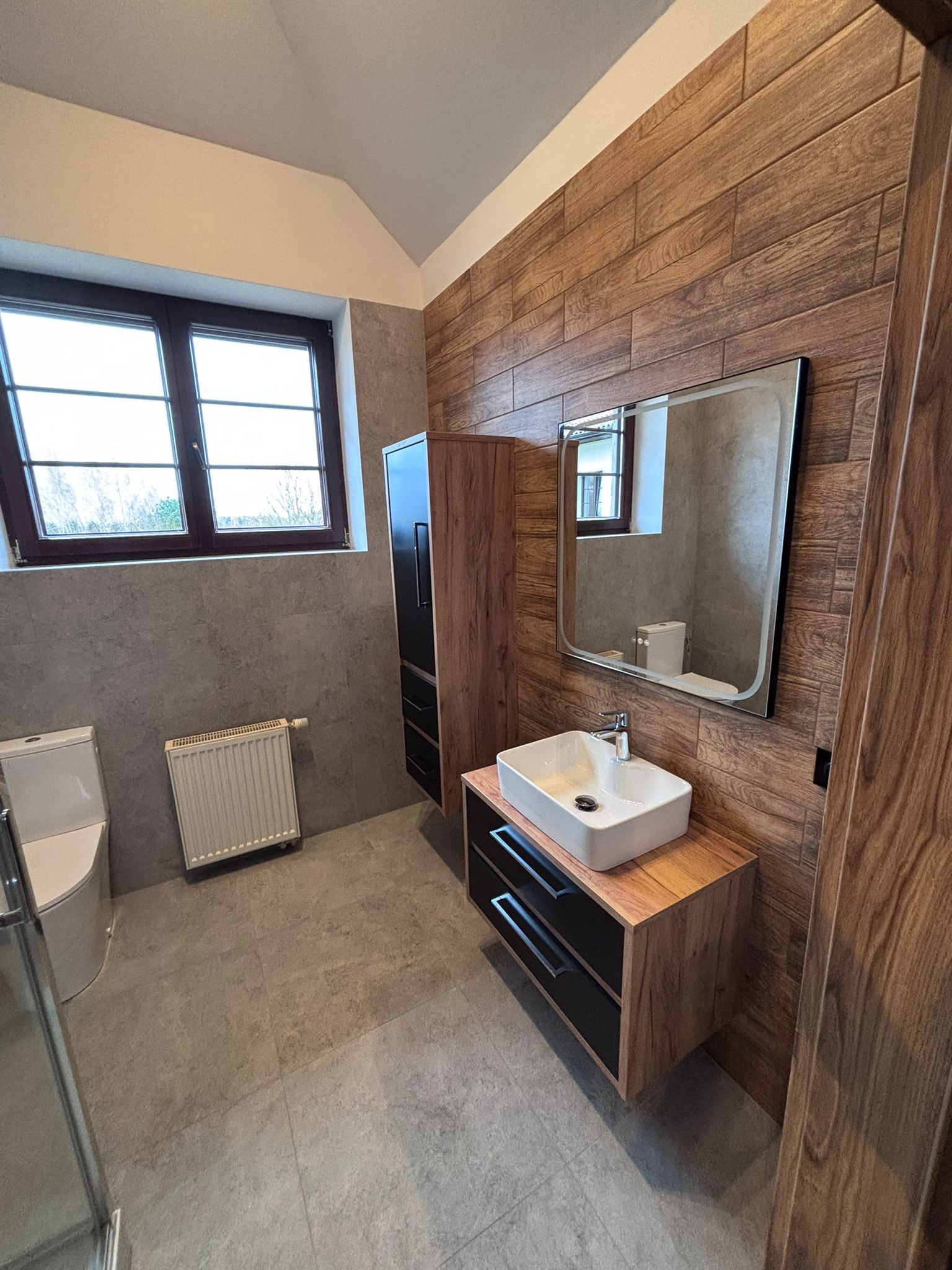 Nowoczesna łazienka z drewnianymi akcentami: ściana z paneli, szafka pod umywalką z czarnymi szufladami, lustro z podświetleniem i grzejnik. Minimalistyczny design.