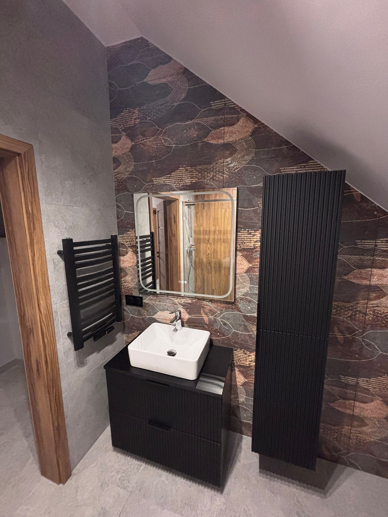 Nowoczesna łazienka na poddaszu z umywalką nablatową, grzejnikiem drabinkowym i szafką w kolorze czarnym. Dekoracyjne płytki na ścianie dodają wnętrzu charakteru.