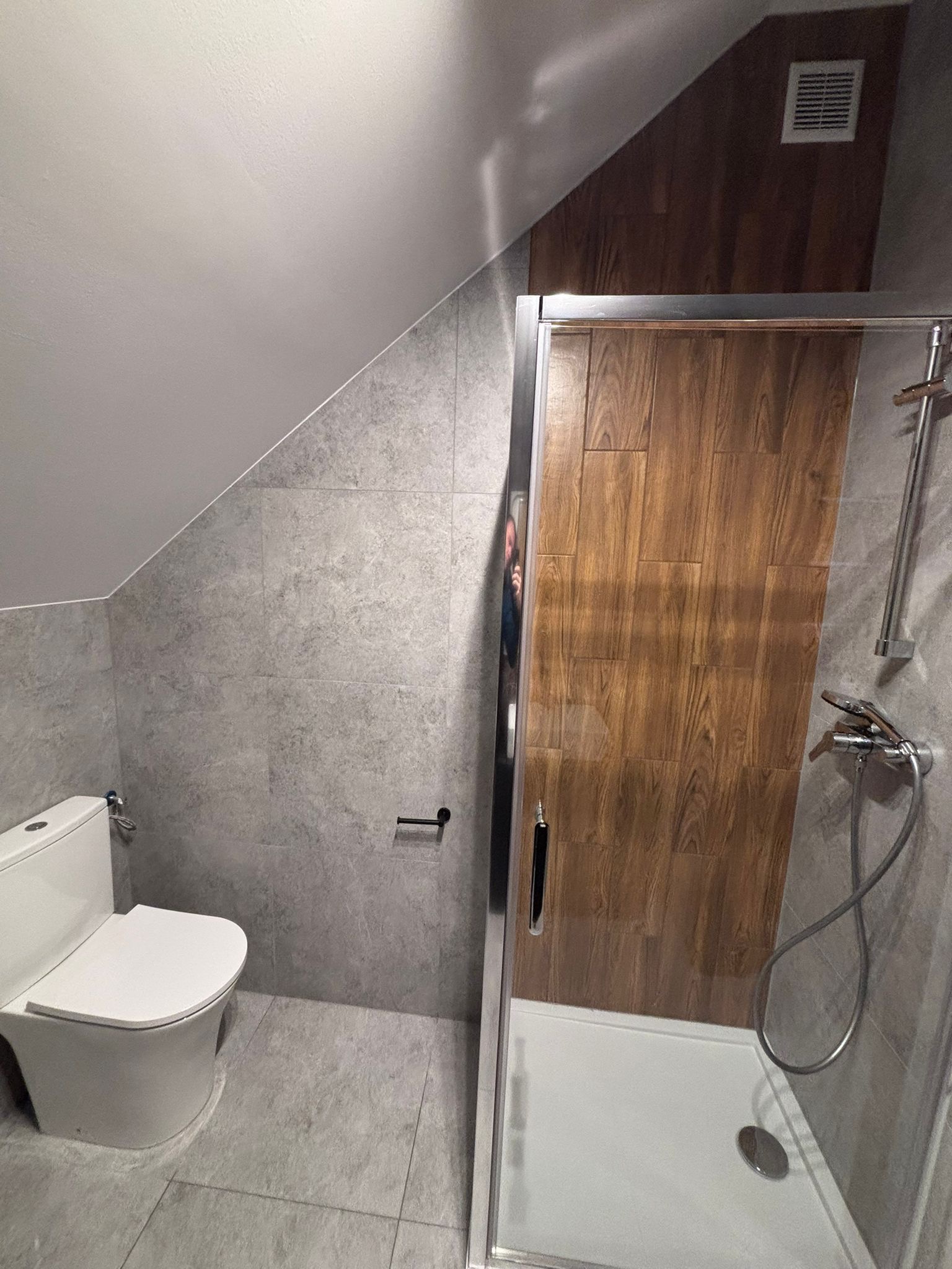 Szara łazienka na poddaszu ze skośnym sufitem, z toaletą i kabiną prysznicową z drewnopodobnymi drzwiami. Widoczna wentylacja.