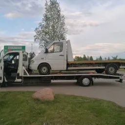 Laweta z białym busem na platformie, zaparkowana na poboczu drogi z zielonym znakiem drogowym wskazującym kierunek na Osiedle Leśne.