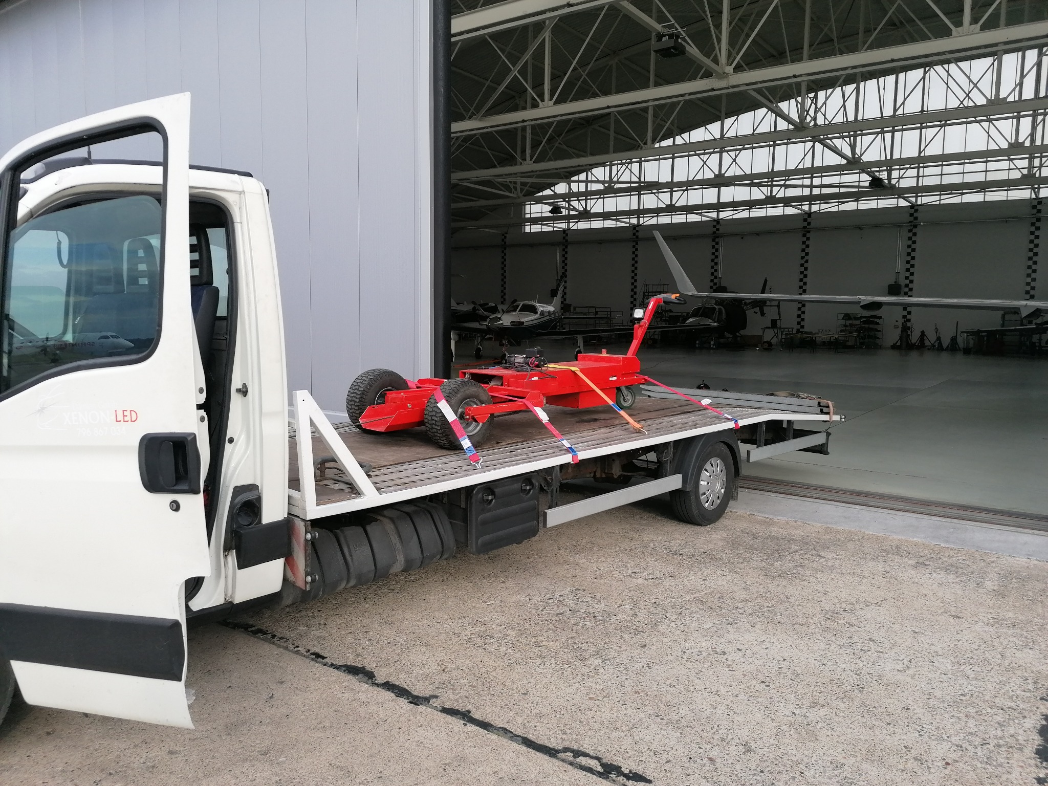 Biała laweta z otwartymi drzwiami kabiny, przewożąca czerwony wózek lotniskowy na tle hangaru z samolotami.
