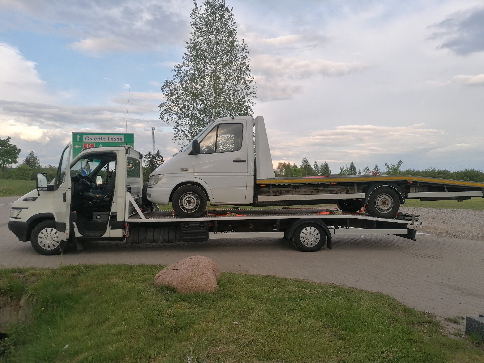 Laweta z białym busem na platformie, zaparkowana na poboczu drogi z zielonym znakiem drogowym wskazującym kierunek na Osiedle Leśne.