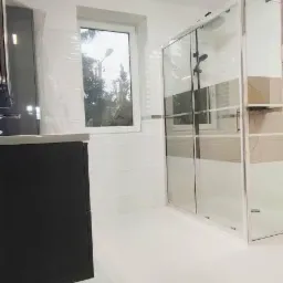 Nowoczesna łazienka z kabiną prysznicową typu walk-in z chromowanymi detalami, czarną szafką pod umywalką i dużym oknem z widokiem na drzewa.