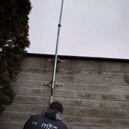 KNM inż. Mateusz Kyrcz - Montaż dwóch kamer na metalowym maszcie przymocowanym do szarej ściany budynku, widok z dołu, pracownik w kurtce z logo firmy widoczny w dolnej części kadru.