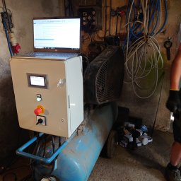 KNM inż. Mateusz Kyrcz - Panel sterowania z ekranem dotykowym i przyciskiem awaryjnym, umieszczony na niebieskim kompresorze, z laptopem z otwartym programem na górze, w tle ściana z kablami i osprzętem elektrycznym.