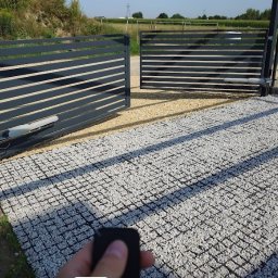 KNM inż. Mateusz Kyrcz - Nowoczesna, automatyczna brama przesuwna z szarymi panelami, widoczny napęd i pilot w dłoni, na podłożu z jasnego kruszywa. Brama otwarta, w tle zieleń.