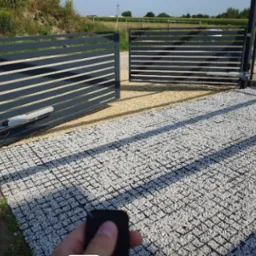 Nowoczesna, automatyczna brama przesuwna z szarymi panelami, widoczny napęd i pilot w dłoni, na podłożu z jasnego kruszywa. Brama otwarta, w tle zieleń.