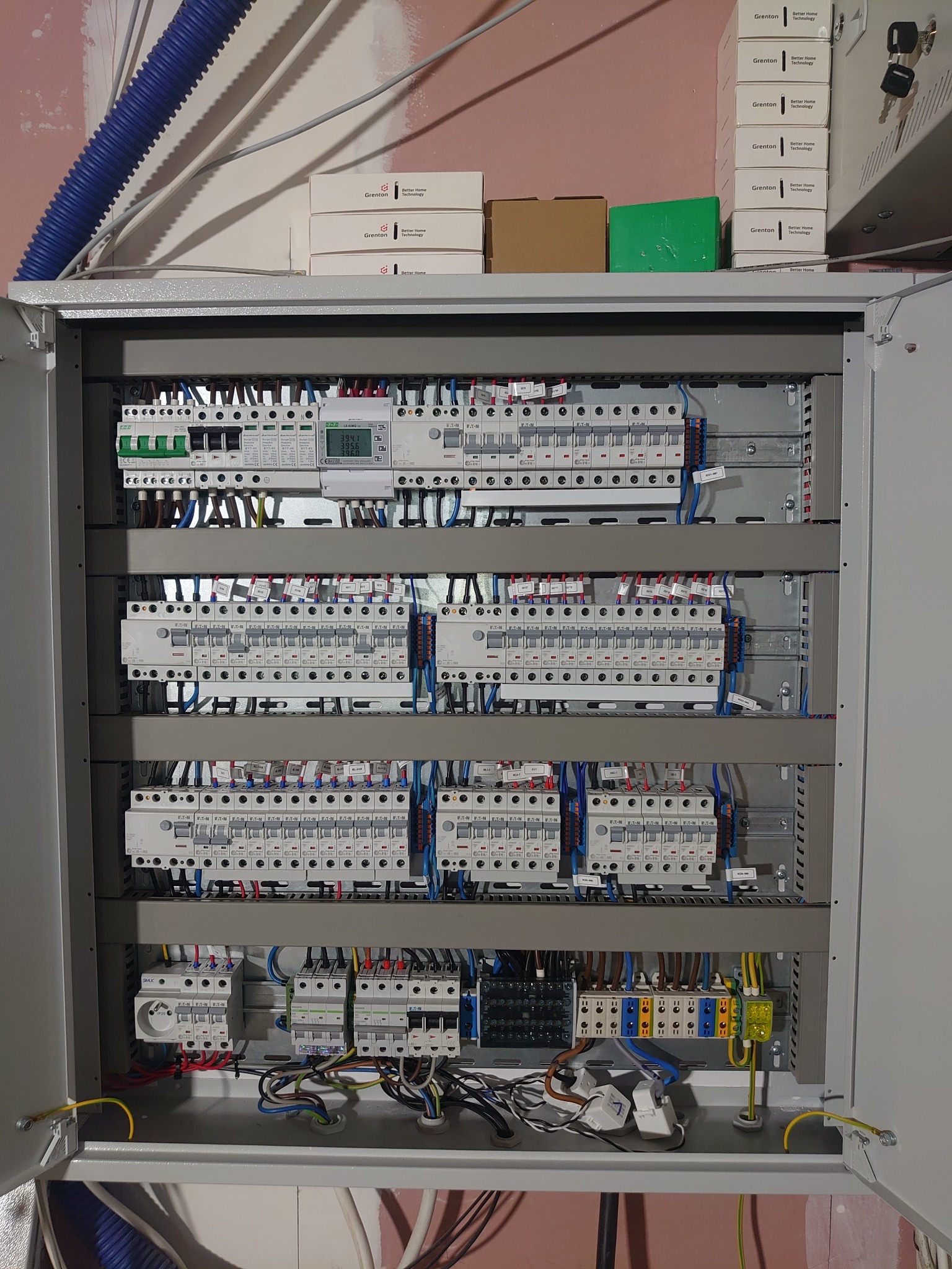 Otwarta szafa rozdzielcza z zainstalowanymi bezpiecznikami, wyłącznikami, przewodami i licznikiem energii elektrycznej; na górze szafy pudełka z logo Grenton Better Home Technology.