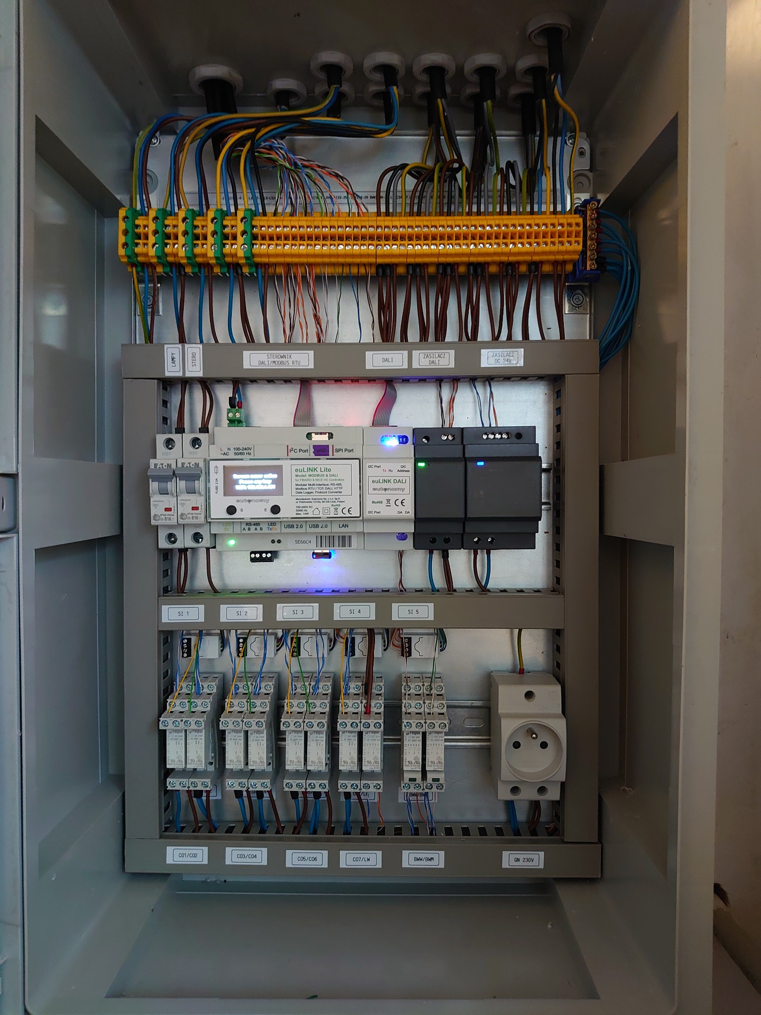 Wnętrze szafy sterowniczej z modułami elektronicznymi, złączkami, przewodami i gniazdem 230V, widoczne diody LED i etykiety opisujące komponenty.