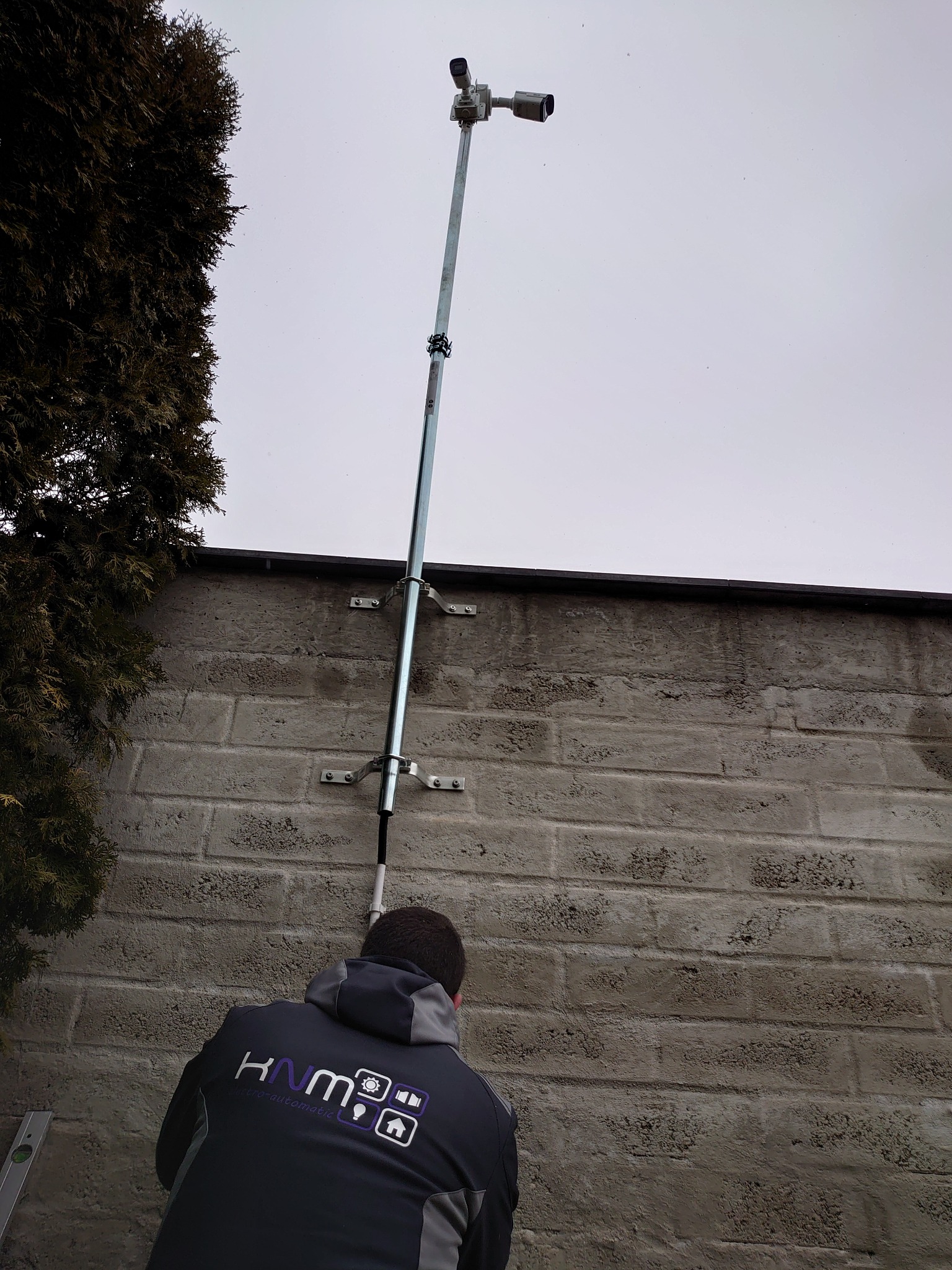 Montaż dwóch kamer na metalowym maszcie przymocowanym do szarej ściany budynku, widok z dołu, pracownik w kurtce z logo firmy widoczny w dolnej części kadru.