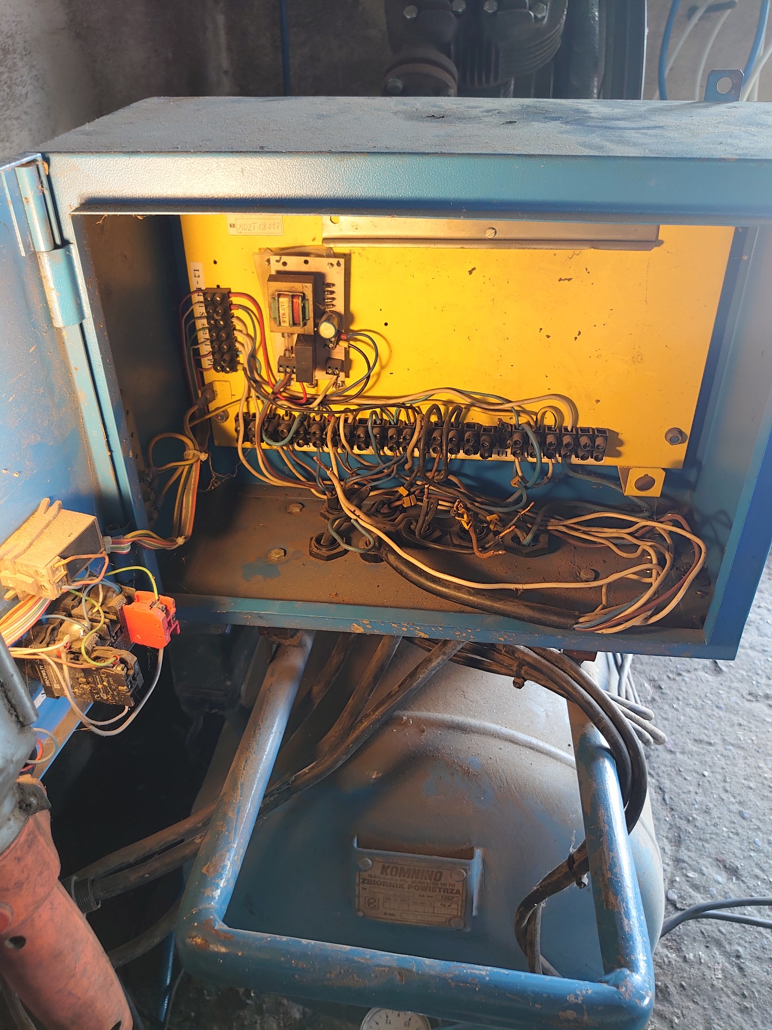 Otwarta skrzynka elektryczna z widocznymi przewodami i elementami sterującymi, zamontowana na kompresorze. Widoczne złącza, bezpieczniki i okablowanie. Całość w niebieskiej obudowie z żółtym tłem...