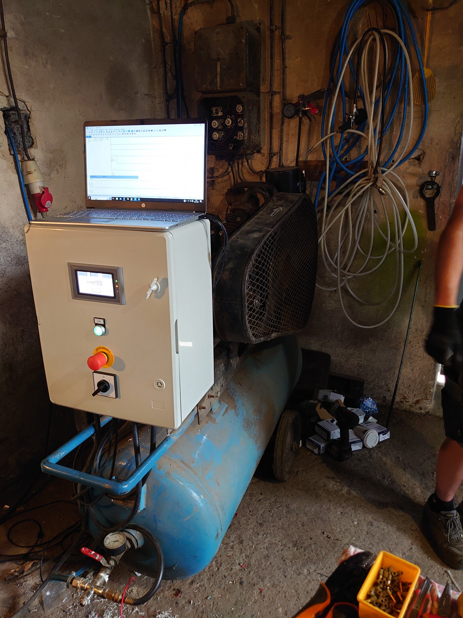 Panel sterowania z ekranem dotykowym i przyciskiem awaryjnym, umieszczony na niebieskim kompresorze, z laptopem z otwartym programem na górze, w tle ściana z kablami i osprzętem elektrycznym.