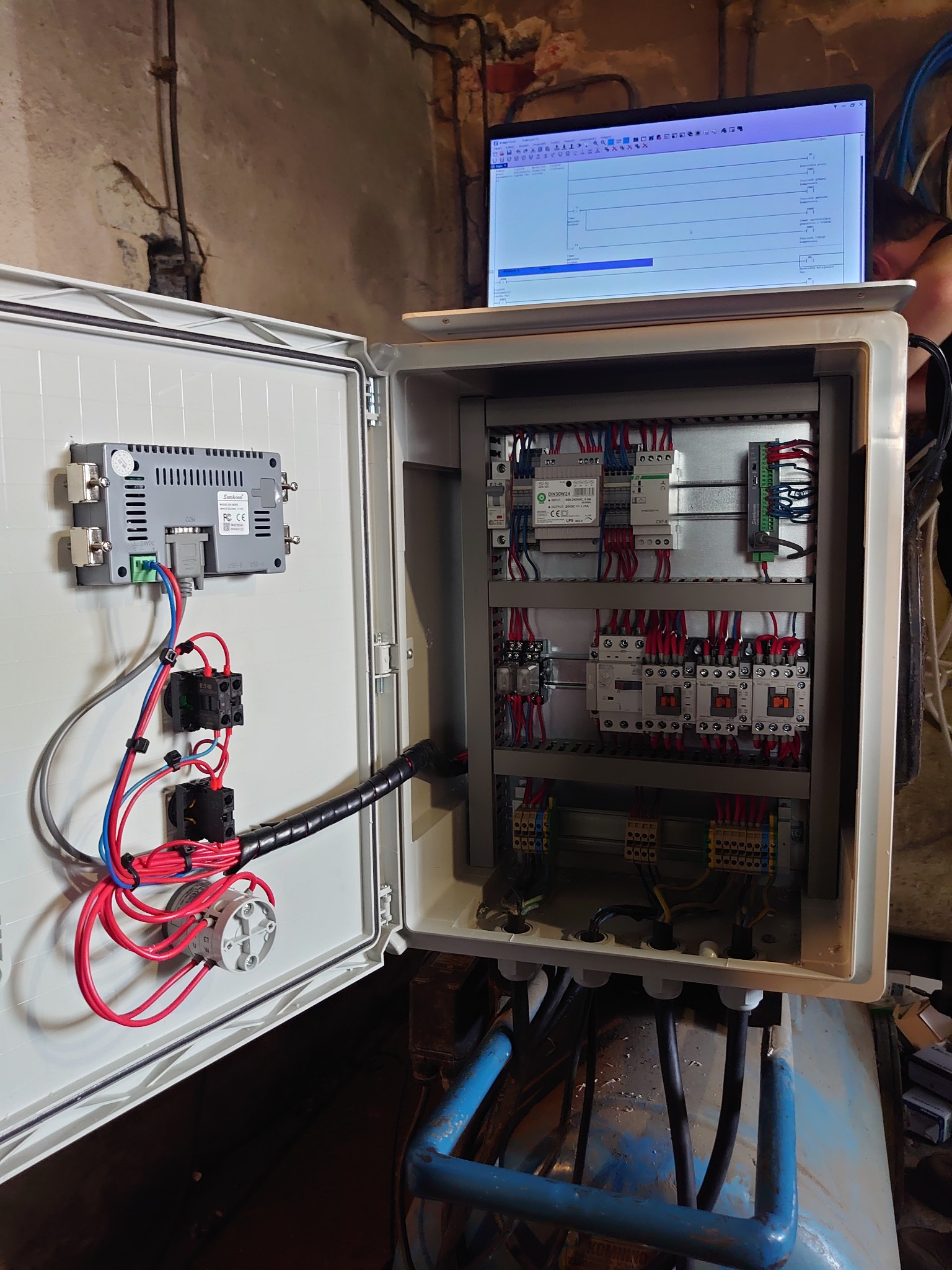 Otwarta szafa sterownicza z widocznymi elementami elektrycznymi, przewodami i modułami, z laptopem na górze wyświetlającym schemat elektryczny, w tle surowa ściana z instalacjami.