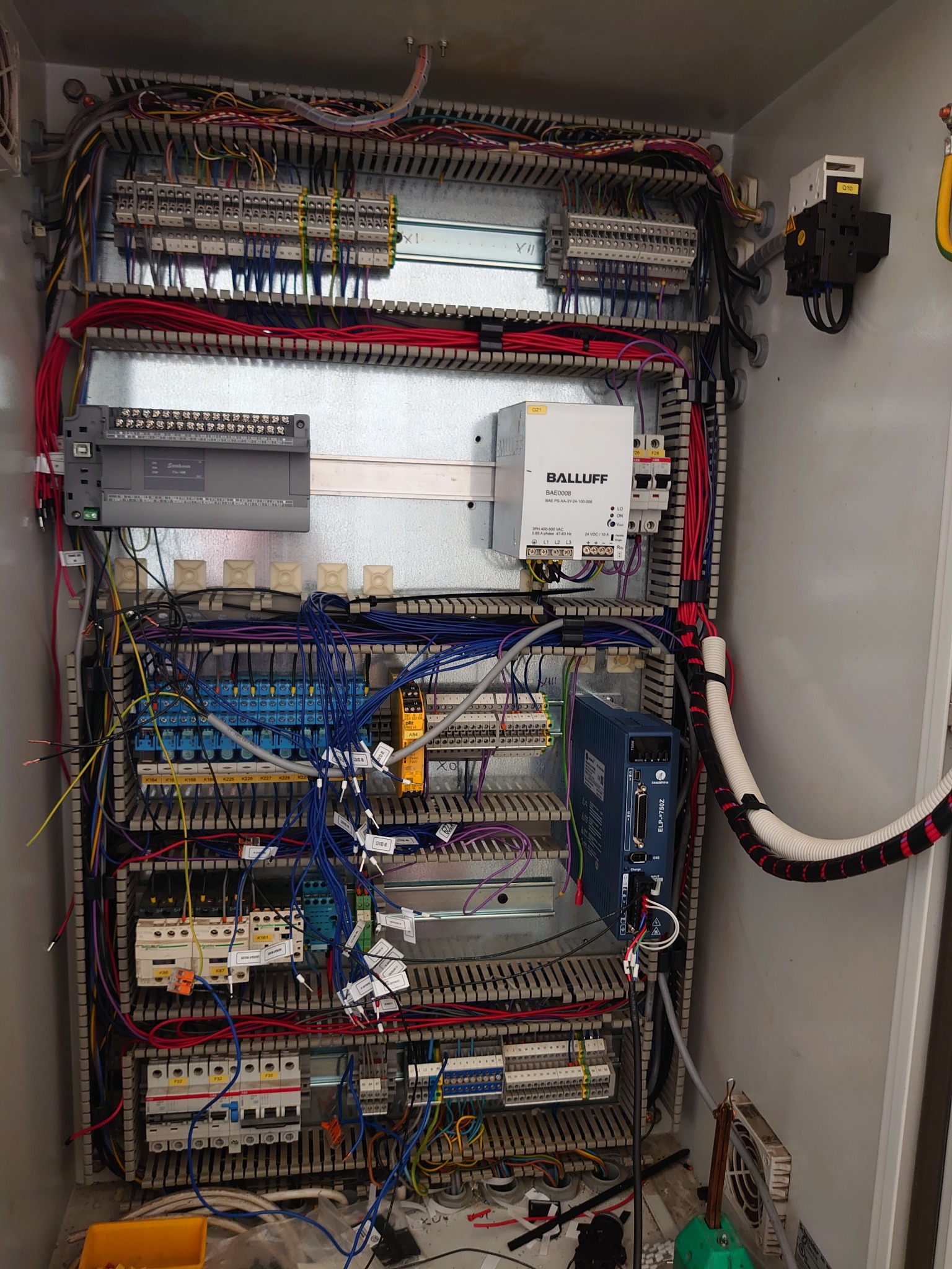 Otwarta szafa sterownicza z licznymi przewodami, modułami i złączkami, widoczne komponenty Balluff i Siemens, przewody w różnych kolorach uporządkowane w listwach, żółta kuweta z elementami na dole...