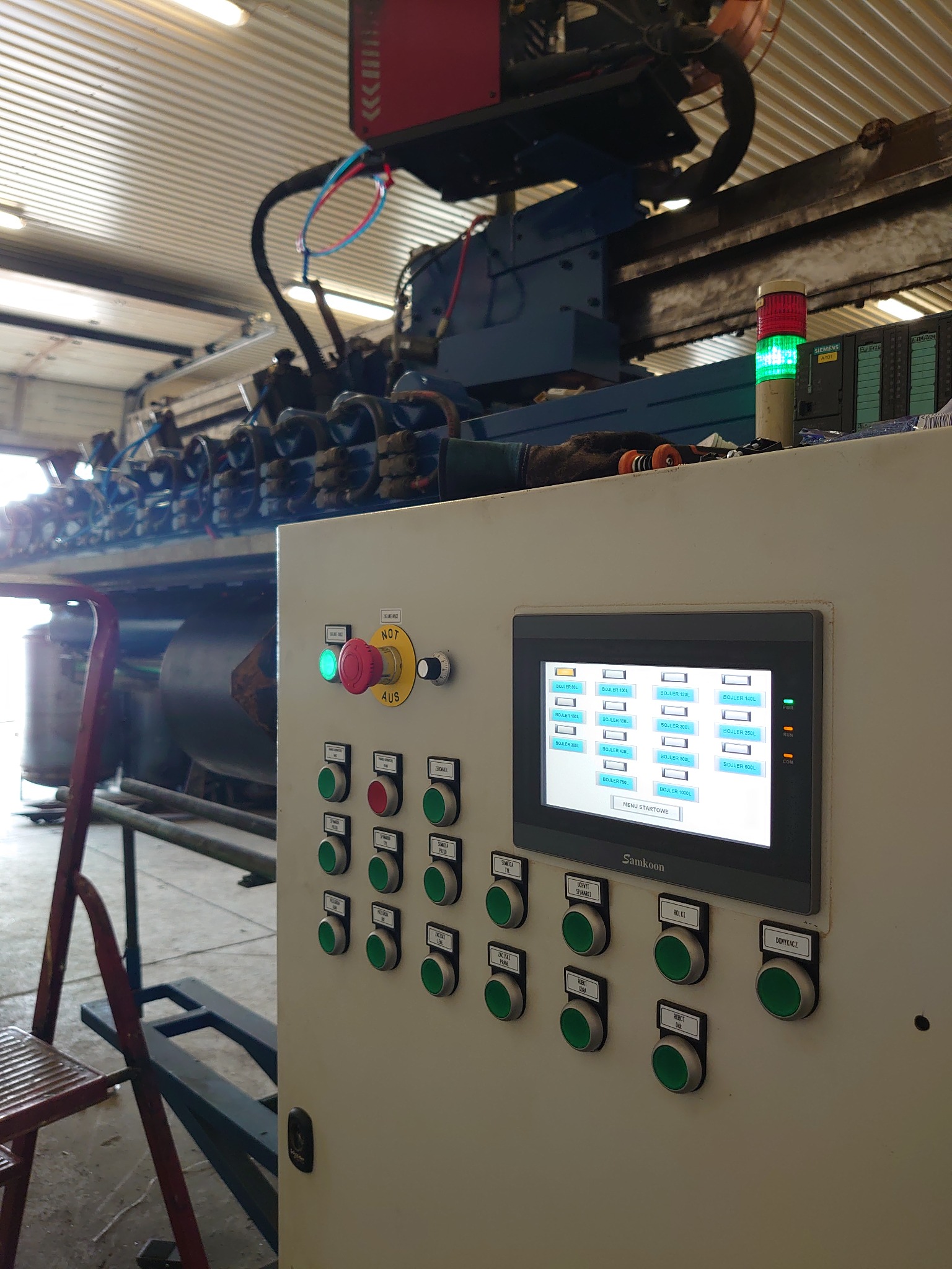 Panel sterowania z ekranem dotykowym i przyciskami, widoczny fragment linii produkcyjnej z rurami i zbiornikami, oświetlenie przemysłowe.
