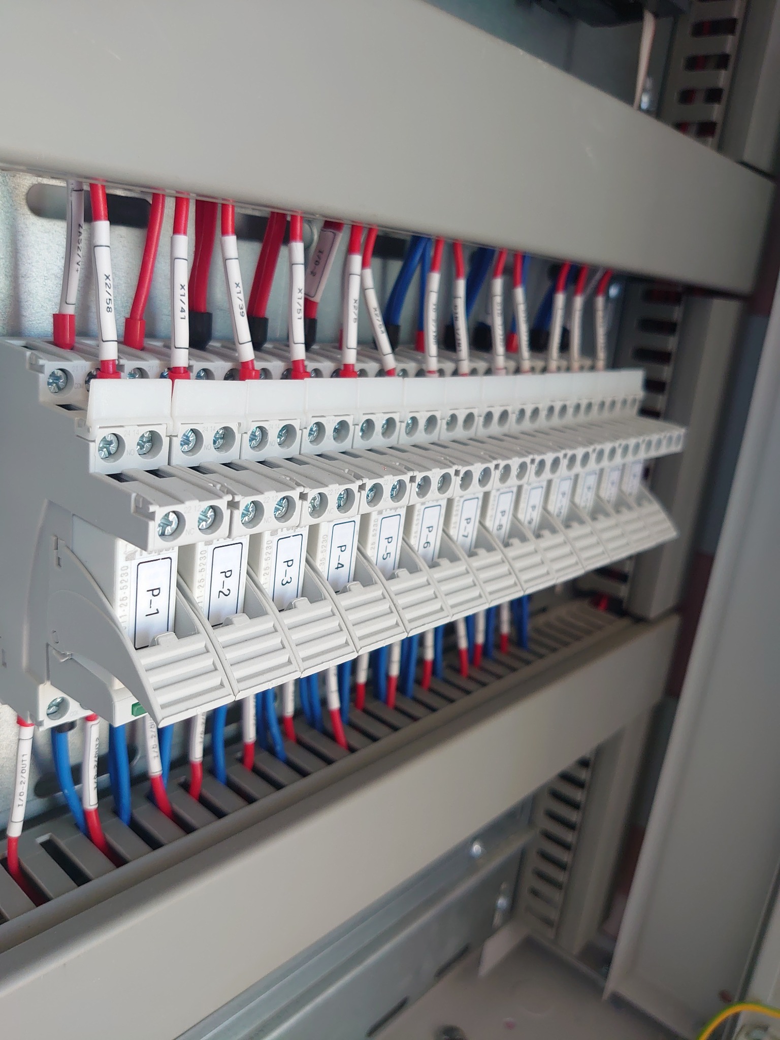 Panel elektryczny z listwami zaciskowymi, oznaczonymi przewodami w kolorach czerwonym i niebieskim oraz etykietami z numerami P-1 do P-7, zamontowany w szarej obudowie.