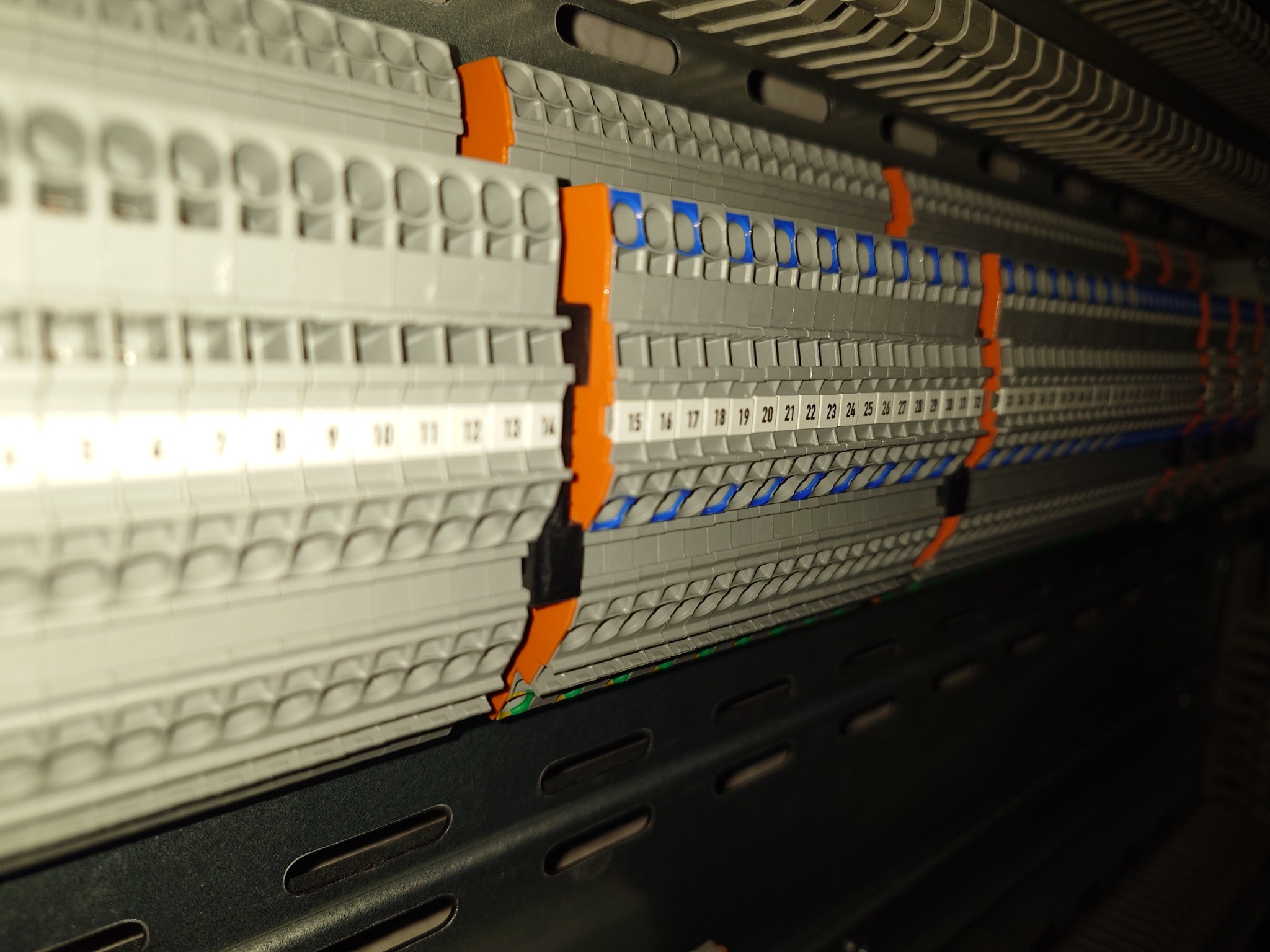 Szyna DIN z listwą zaciskową, widoczne ponumerowane zaciski z niebieskimi i pomarańczowymi znacznikami, zamontowana w szafie elektrycznej.