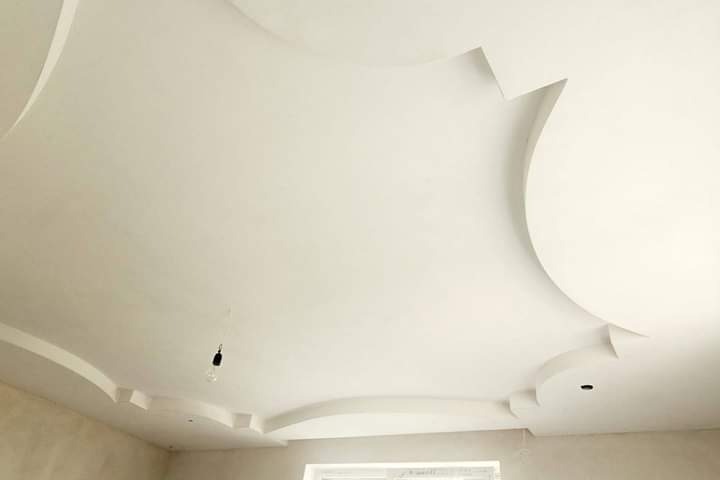 Wykończony sufit podwieszany z gładzią i dekoracyjnymi elementami w kształcie łuków i kwadratów, widoczna żarówka na kablu.