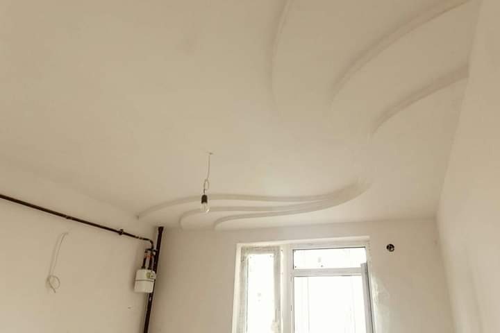 Biały sufit z dekoracyjnymi, gipsowymi listwami w kształcie fal, wisząca żarówka na kablu, instalacja gazowa przy ścianie, okno PCV.