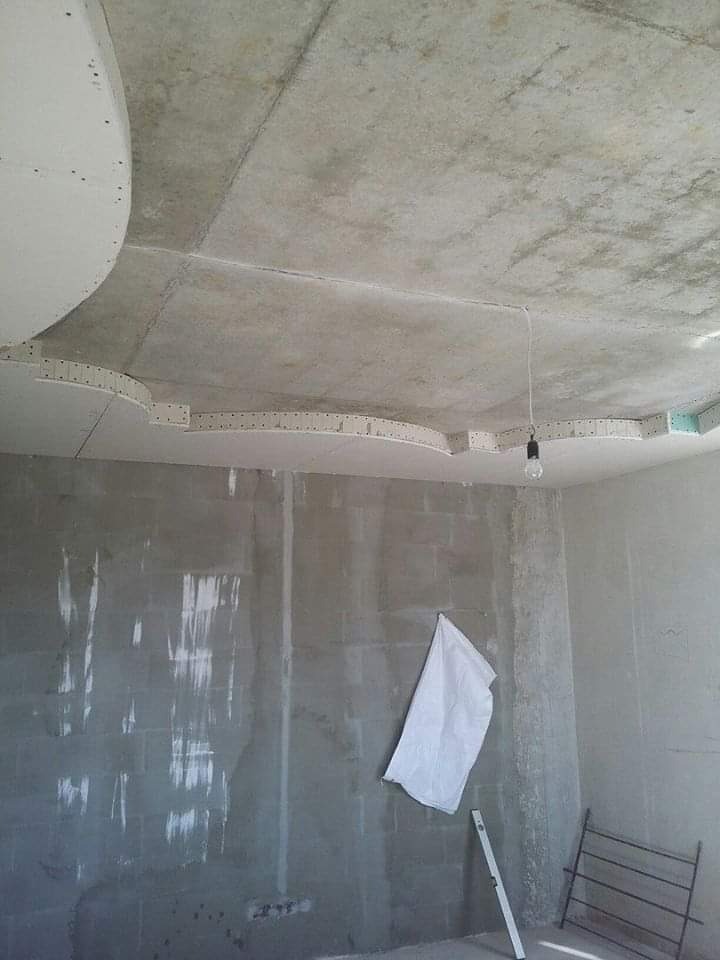 Surowy betonowy sufit z elementami podwieszanego sufitu gipsowo-kartonowego w trakcie montażu, widoczna goła żarówka i ściany z surowego betonu, przygotowywane do dalszych prac wykończeniowych.