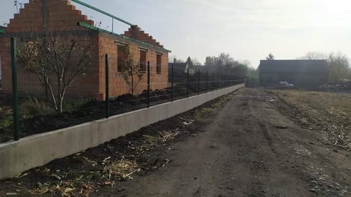 Nowoczesne ogrodzenie panelowe z podmurówką betonową na tle surowego budynku z cegły i polnej drogi.