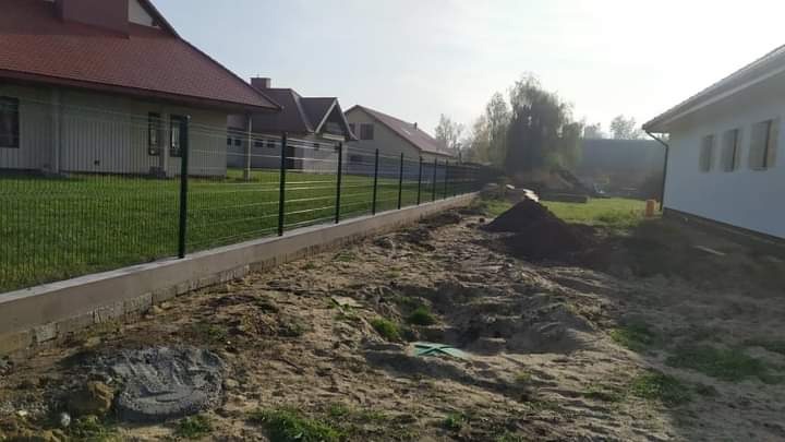 Montaż ogrodzenia panelowego na betonowej podmurówce, widoczne domy w tle i przygotowany teren pod trawnik.