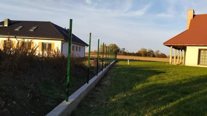 Wkopane, zielone słupki ogrodzeniowe w betonowej podmurówce, widoczne na tle domów jednorodzinnych i trawnika w słoneczny dzień.