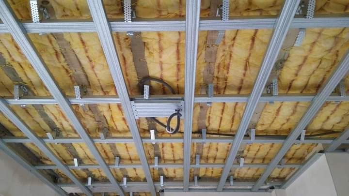 Montaż izolacji z wełny mineralnej pomiędzy metalowym stelażem sufitu podwieszanego, widoczne przewody elektryczne.