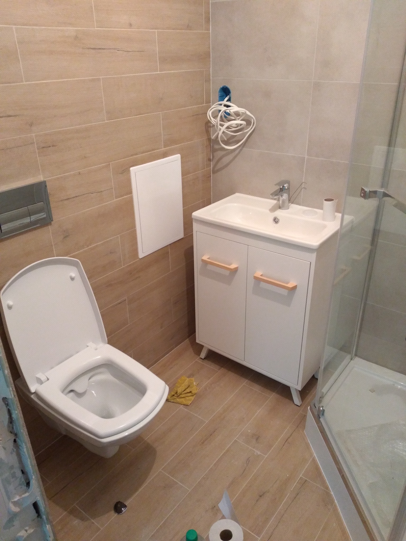 Ukończona łazienka z beżowymi płytkami imitującymi drewno, białą toaletą z otwartą klapą, białą szafką pod umywalkę z drewnianymi uchwytami i kabiną prysznicową z przezroczystego szkła; widoczne...