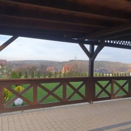 F.H.U. TreeBiel Piotr Bielak - Zadaszony taras z drewnianą balustradą w kształcie krzyży, widok na zielony trawnik, domy i wzgórza, na balustradzie siedzi czarny kot.