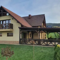 Dom jednorodzinny z brązową, drewnianą balustradą balkonową i zadaszonym tarasem z drewnianą konstrukcją, otoczony zielonym trawnikiem i młodymi drzewkami.