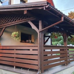 F.H.U. TreeBiel Piotr Bielak - Zadaszona konstrukcja z drewnianą balustradą, prawdopodobnie na tarasie, z wiszącymi doniczkami i widocznym oknem domu w tle.