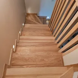 Nowoczesne schody drewniane z oświetleniem punktowym i ażurową balustradą, widok z góry, ukazujący naturalny rysunek drewna i minimalistyczny design.