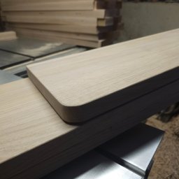 F.H.U. TreeBiel Piotr Bielak - Obróbka drewna: Frezowana deska z zaokrąglonym rogiem na stole warsztatowym, w tle stos desek. Widoczne ślady obróbki.