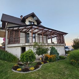 F.H.U. TreeBiel Piotr Bielak - Dom z zadaszonym tarasem z drewnianą konstrukcją i balustradą. Widoczne elementy ogrodu i oświetlenie. Ujęcie z dołu, w tle błękitne niebo.