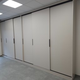 F.H.U. TreeBiel Piotr Bielak - Długa, beżowa szafa wnękowa z przesuwnymi drzwiami i czarnymi uchwytami, dopasowana do szarej podłogi z płytek. Minimalistyczny design, oświetlenie sufitowe.
