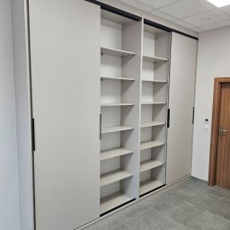 F.H.U. TreeBiel Piotr Bielak - Biała szafa wnękowa z przesuwnymi drzwiami i czarnymi uchwytami, widoczne półki wewnątrz, szara podłoga z płytek, minimalistyczny design.
